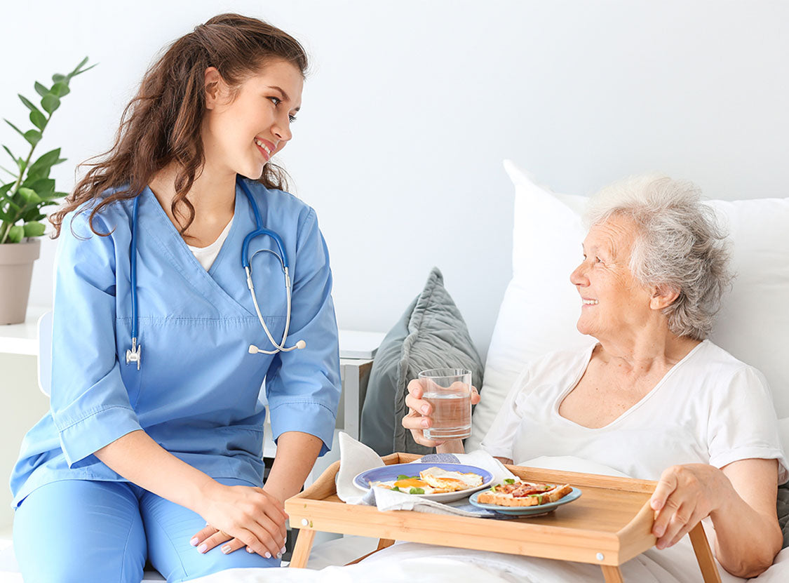 Nurse in blue scrubs serving food to an elderly patient in bed.